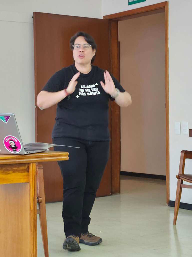 Mujer vestida de negro junto a un escritorio hablando hacia el frente, en un salón.