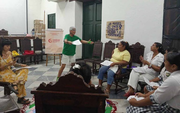 Mujeres reunidas en un salón, sentadas mientras una de pie se dirige a ellas.