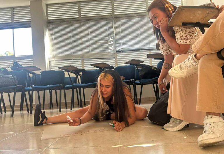 Una mujer sentada en el piso escribiendo en una cartelera, mientras otras observan sentadas desde sillas escritorio.