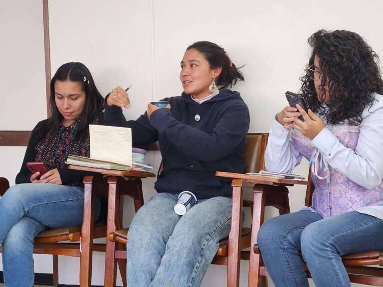 Tres mujeres sentadas en sillas escritorio; la del centro habla hacia el frente.