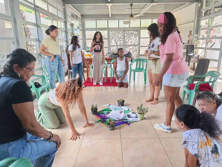 Grupo de mujeres en un salón en actividad que las reúne hacia el centro del espacio donde hay algunos elementos dispuestos.